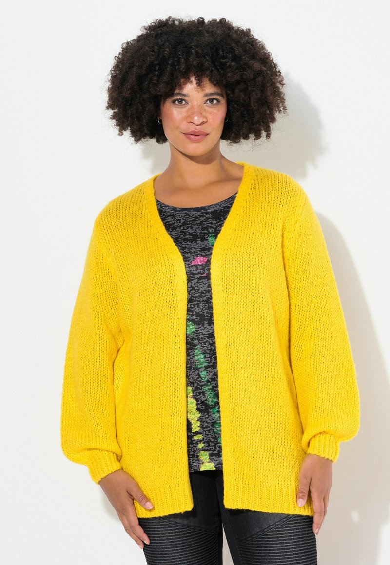 Cardigan giallo a maglia con vestibilità ampia, davanti aperto e polsini a coste. Indossato sopra una maglietta nera a fantasia, con tonalità di verde e rosa.