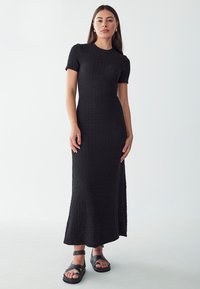 Schwarzes gestricktes Maxikleid mit strukturiertem Finish, kurzen Ärmeln und ausgestelltem Saum. Kombiniert mit schwarzen, sandalettenartigen Schuhen. Einfache und moderne Gestaltung.