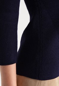 Pull en tricot bleu marine avec une texture côtelée et des manches trois-quarts, présentant des motifs de couture complexes sur les côtés.