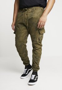 Kamo cargo bukser i olivengrøn, med snor i taljen, sidelommer og en tætsiddende pasform. Sammen med sorte sneakers.
