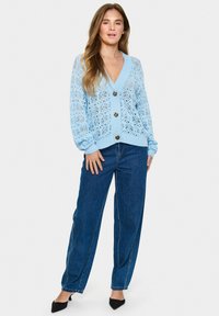 Ljusblå stickad cardigan med spetsmönster, ribbade ärmslut och fem guldknappar, tillsammans med högmidjade jeans i denim och svarta klackar.