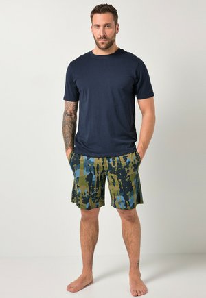 Mand stående barfodet iført navyblå T-shirt og grønne, blå og sorte abstrakte camo-shorts med hænderne i lommerne, mod en ensfarvet baggrund.