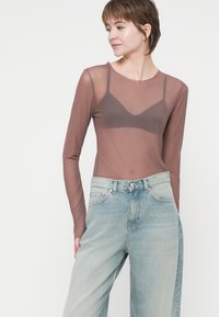 Dolga, prosojna rjava bluza, v kombinaciji s svetlim, visokim jeansom. Bluza ima kroj z okroglim izrezom in subtilen dizajn, podoben modrčku.