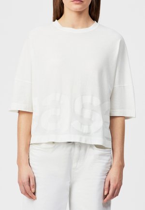 Mujer con una camisa blanca de manga corta de gran tamaño con texto grande y tenue, y pantalones blancos, de pie contra un fondo liso.