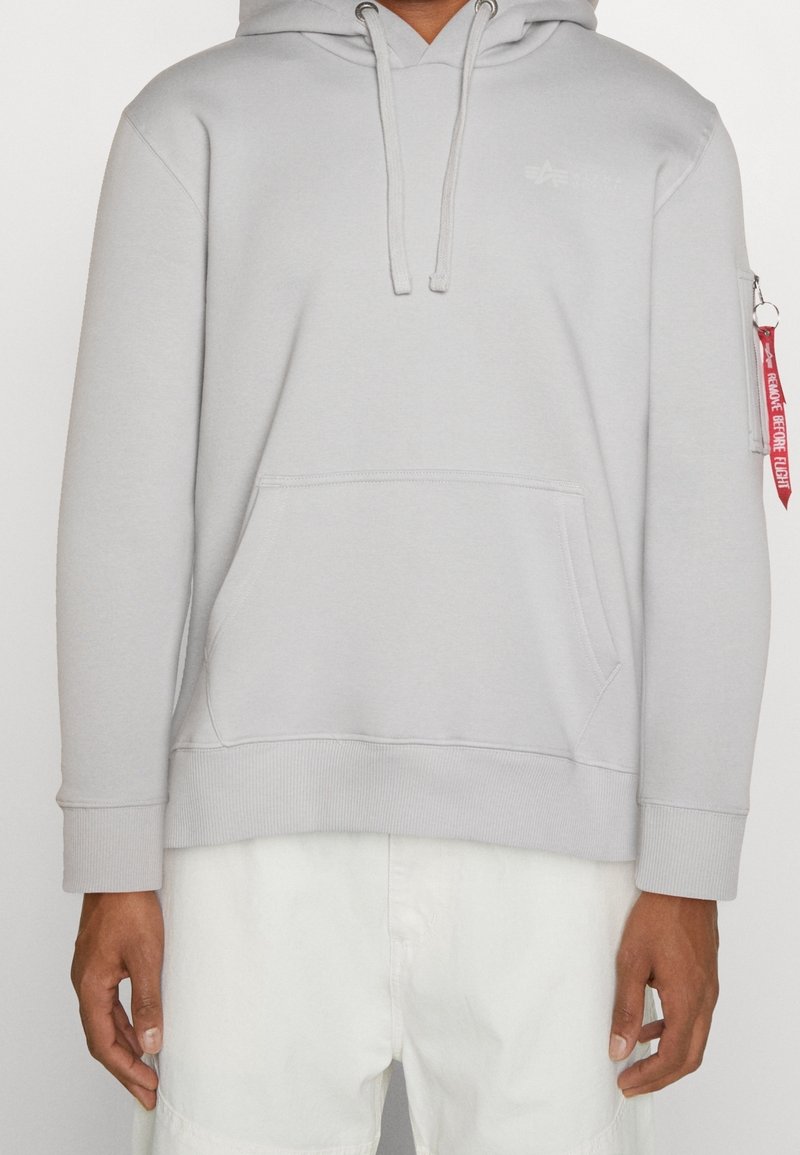 Sudadera gris claro con un bolsillo frontal, cordones y puños acanalados. Presenta un pequeño logo en el pecho y una etiqueta roja en la manga.