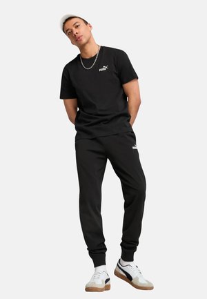 Zwarte T-shirt met korte mouwen en een klein wit logo, gecombineerd met zwarte joggers met een logo. Witte en beige sneakers maken de look compleet.