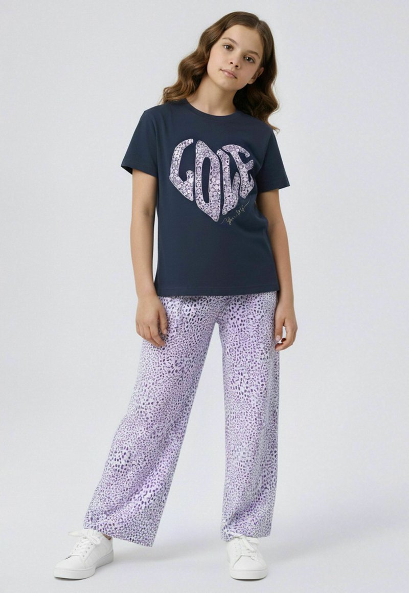 Jeune fille debout portant un t-shirt bleu marine avec le texte "LOVE" et un pantalon ample lilas à imprimé léopard, ainsi que des baskets blanches.