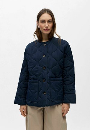 Femme portant une veste matelassée bleu marine à boutons avec de grandes poches avant et un pantalon beige, debout devant un fond blanc uni.