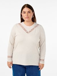 Top beige a maniche lunghe con dettagli in pizzo sul collo a V e sui polsini, texture morbida, vestibilità rilassata, abbinato a jeans blu.
