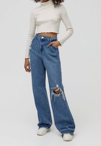 Valkoinen ribbattu lyhyt turtlenekte-pullover, jossa on aaltoilevat reunat, yhdistettynä korkeavyötäröisiin sinisiin farkkulleihin, joissa on revityt polvet.