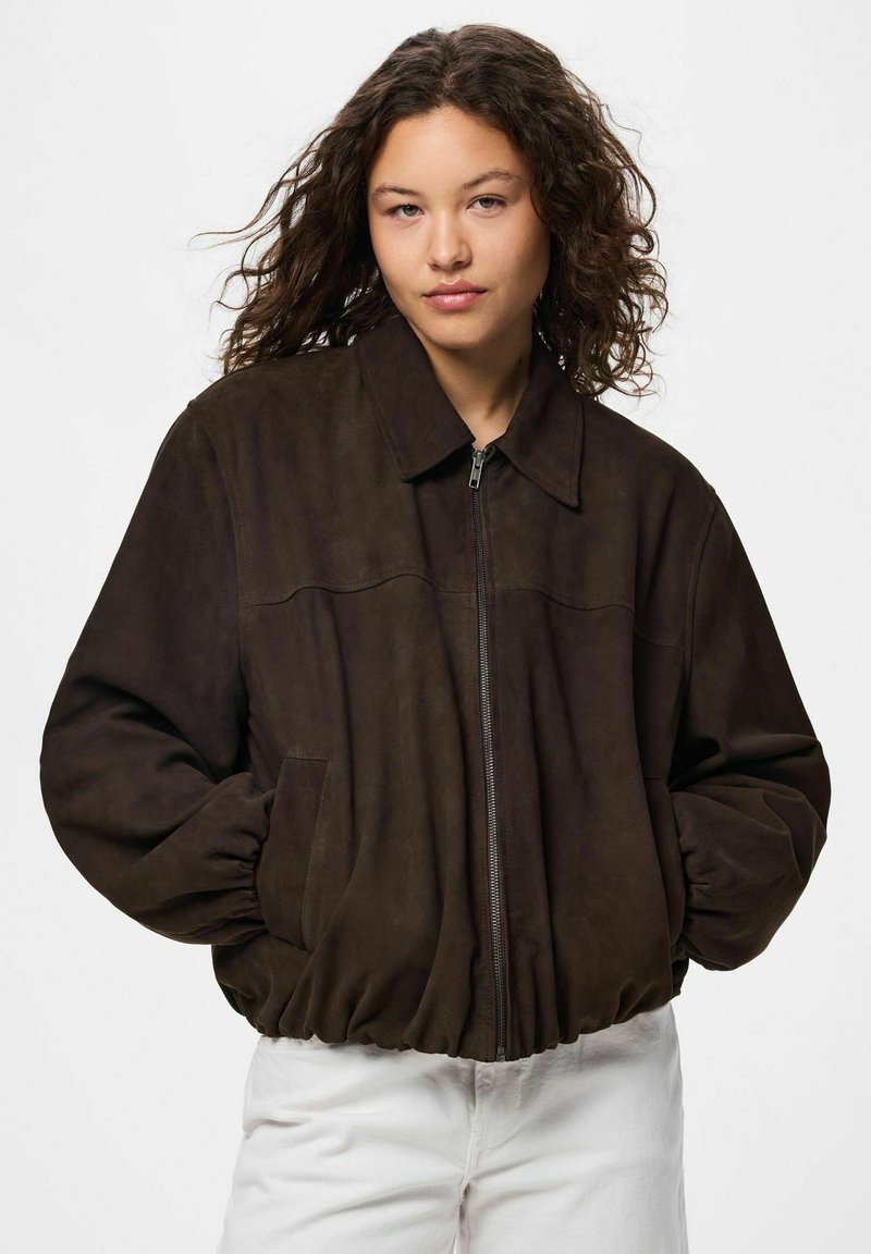 Jeune femme aux cheveux bouclés portant une veste zippée marron foncé avec des poches, debout devant un fond clair uni.