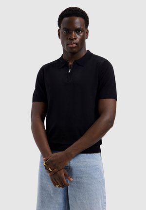 Junger Mann, der ein schwarzes Polo mit kurzen Ärmeln und Reißverschluss, hellblaue Jeans und mehrere goldene Ringe an den Fingern trägt, vor schlichtem grauem Hintergrund.