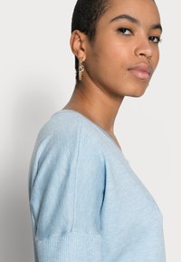 Lyseblå V-hals sweater lavet af fint strikket stof med rippede ærmekanter. Modellen bærer guldøreringe for ekstra detaljer.