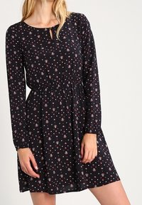 Svart långärmad klänning med blommönster, med insydd midja och detalj med nyckelhål i halsringningen. Lättviktigt tyg med en flödande silhuett.