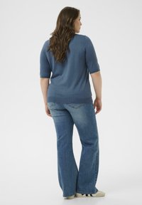 Blaues gestricktes Kurzarm-Oberteil, kombiniert mit hellblauen ausgestellten Jeans. Sanfte Textur, lockere Passform und minimale Details; die Schuhe sind cremefarben mit schwarzen Streifen.
