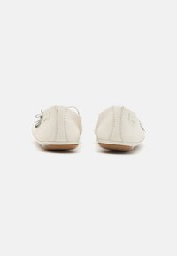 Los zapatos de cuero blanco tipo slip-on presentan una textura suave, forma redondeada, suelas delgadas y un adorno de moño gris decorativo en la parte frontal.