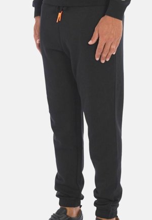 Uomo che indossa pantaloni jogger neri con polsini elastici e coulisse arancione, in piedi con le mani rilassate lungo i fianchi.
