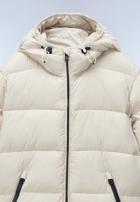 Beige pufferjack met een capuchon, voorzien van gewatteerde vulling, een zwarte rits, en verstelbare trekkoorden op de capuchon en de zoom.