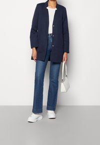 Abrigo largo azul marino con cuello puntiagudo y cierre de botones, usado sobre una camiseta blanca de canalé y jeans acampanados azules, combinado con zapatillas blancas.