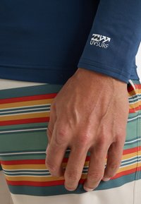 Marinblå långärmad rash guard med UV-skydd och logga på ärmen; handen vilar på färgglada randiga badshorts med gröna, röda, gula och blå linjer.