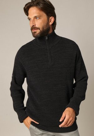 Homme barbu portant un pull à col zippé sombre à texture et un pantalon clair, avec une main dans sa poche, regardant à gauche.