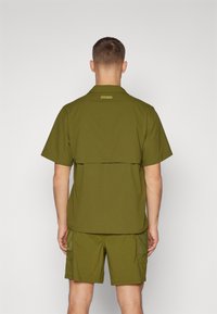 Oliwkowa koszula z krótkim rękawem z tylną klapą, w zestawie z dopasowanymi szortami posiadającymi siatkową kieszeń i teksturowaną tkaninę.