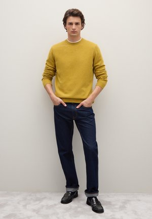 Senfgelber Strickpullover mit Rundhalsausschnitt, kombiniert mit dunkelblauen Jeans und schwarzen Loafers, steht vor einem neutralen Hintergrund.