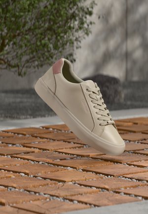 Baskets de couleur crème avec languette rose au talon, lacets beige et empeigne lisse, suspendues au-dessus d'un sol en dalles de bois avec des espaces remplis de gravier en extérieur.