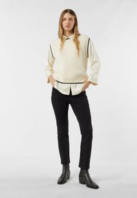 Kremowy bezrękawnik z dzianiny z czarnym wykończeniem, noszony na jasnej koszuli; zestawiony z czarnymi wąskimi spodniami i botkami za kostkę.