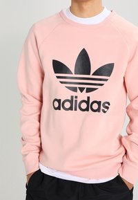 Rosa sweatshirt i bomullsblandning med en svart Adidas-logotyp och tre ränder över bröstet. Ribbstickade muddar och fåll.