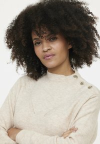 Pull en maille beige avec un col montant, présentant un design texturé et trois boutons décoratifs à l'épaule. Tissu doux et confortable.