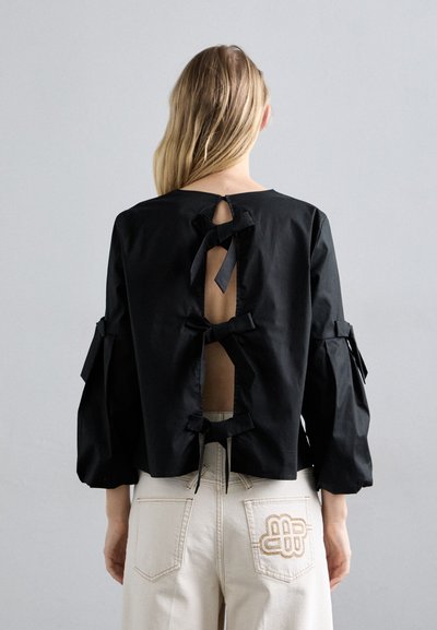 Blouse noire courte avec un col rond, des manches longues et plusieurs accents en ruban à l'arrière, laissant la peau visible à travers des découpes.