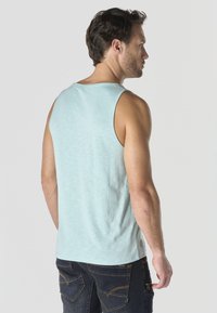 Hombre con camiseta sin mangas azul claro y vaqueros oscuros, de perfil con la cabeza girada hacia la derecha contra un fondo liso.