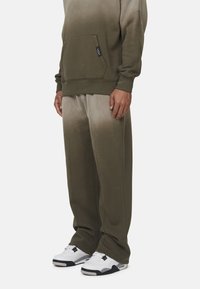 Olijfgroene hoodie met een voorke pocket, gecombineerd met bijpassende taps toelopende sweatpants met een ombre-effect. Draagt met witte sportschoenen.