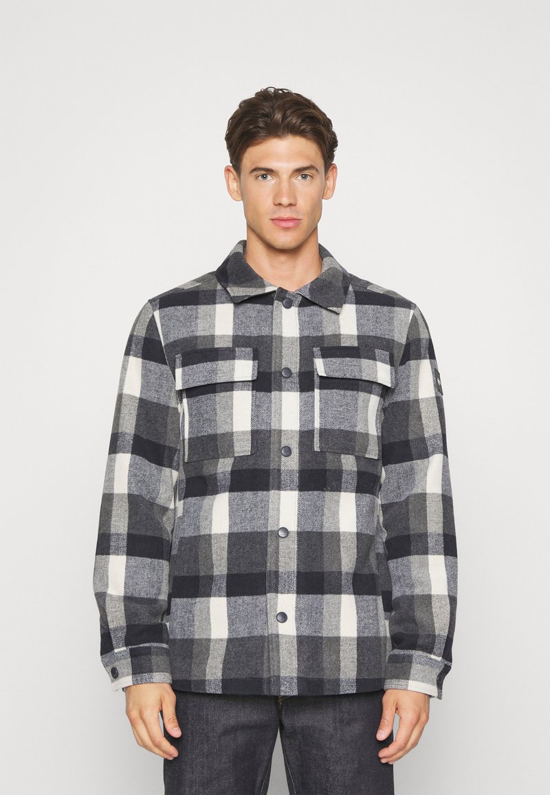 Veste en flanelle à carreaux dans les tons gris, noir et blanc. Comprend deux poches poitrine, une fermeture à boutons et un col classique. Texture douce.