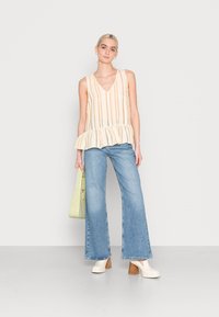 Top de peplum às riscas em bege e creme, design em forma de V; combinado com jeans de perna larga em azul claro; modelo segura uma bolsa verde; usa sapatos de salto em off-white.