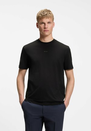 Lyshåret mand står med hænderne i lommerne iført sort kortærmet BOSS T-shirt og marineblå bukser mod en ensfarvet hvid baggrund.