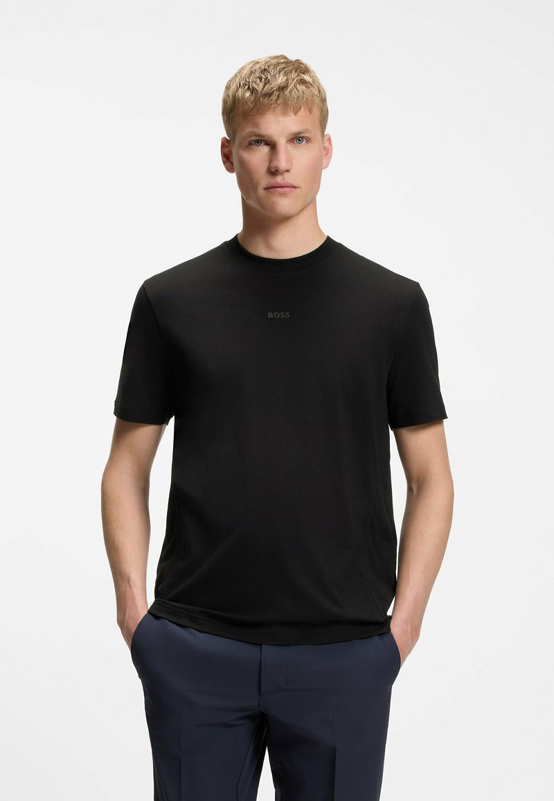 Blonder Mann steht mit den Händen in den Taschen, trägt ein schwarzes kurzärmeliges BOSS T-Shirt und marineblaue Hose vor weißem Hintergrund.