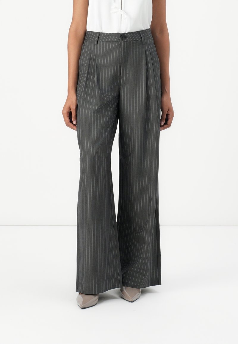 Personne portant un pantalon large gris à fines rayures avec des plis à l'avant et des chaussures beiges à bout pointu, debout devant un fond blanc.