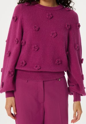 Kobieta ubrana w fioletowy, dziergany sweter z trójwymiarowymi kwiatowymi wzorami, zestawiony z pasującymi fioletowymi, wysokim stanem spodniami.