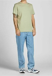 Jasnozielona koszulka z krótkim rękawem o luźnym kroju, zestawiona z jasnoniebieskimi prostymi dżinsami. Buty sportowe mają białe i granatowe akcenty.