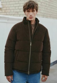 Brun pufferjakke lavet af blød kordfløjl med høj krave, lynlåslukning foran og to sidelommer. Båret over en lys sweater.