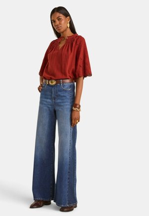 Femme portant un chemisier texturé rouge rouille à manches larges, un jean bleu taille haute à jambes larges, une ceinture marron et des chaussures marron, posant sur fond blanc.