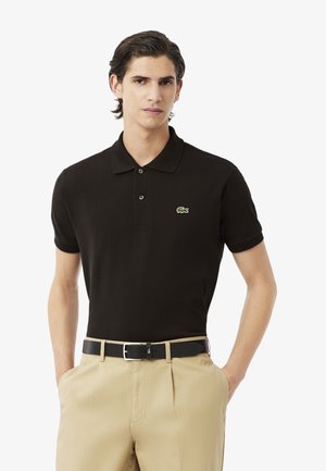 Polo negro de algodón con mangas cortas, que presenta un logotipo verde bordado en el pecho. Se lleva con pantalones beiges y un cinturón negro.