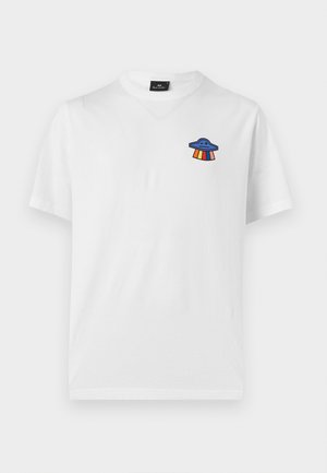 Ung mand står med hænderne i lommerne iført hvid T-shirt med lille farverig UFO-grafik, grå joggingbukser og hvide sneakers på ensfarvet baggrund.