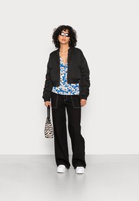 Casaco bomber curto preto sobre uma blusa floral azul, combinado com calças largas pretas e ténis brancos, segurando uma mala às riscas.