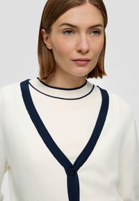 Crème V-hals cardigan met marineblauw accent, voorzien van een knoopsluiting en zachte textuur. Benadrukt contrasterende kleuren en strakke lijnen.
