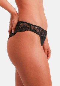 Czarny koronkowy bikini z falistymi brzegami, półprzezroczystym przednim panelem i gładkim tyłem, podkreślający delikatne kwiatowe wzory.