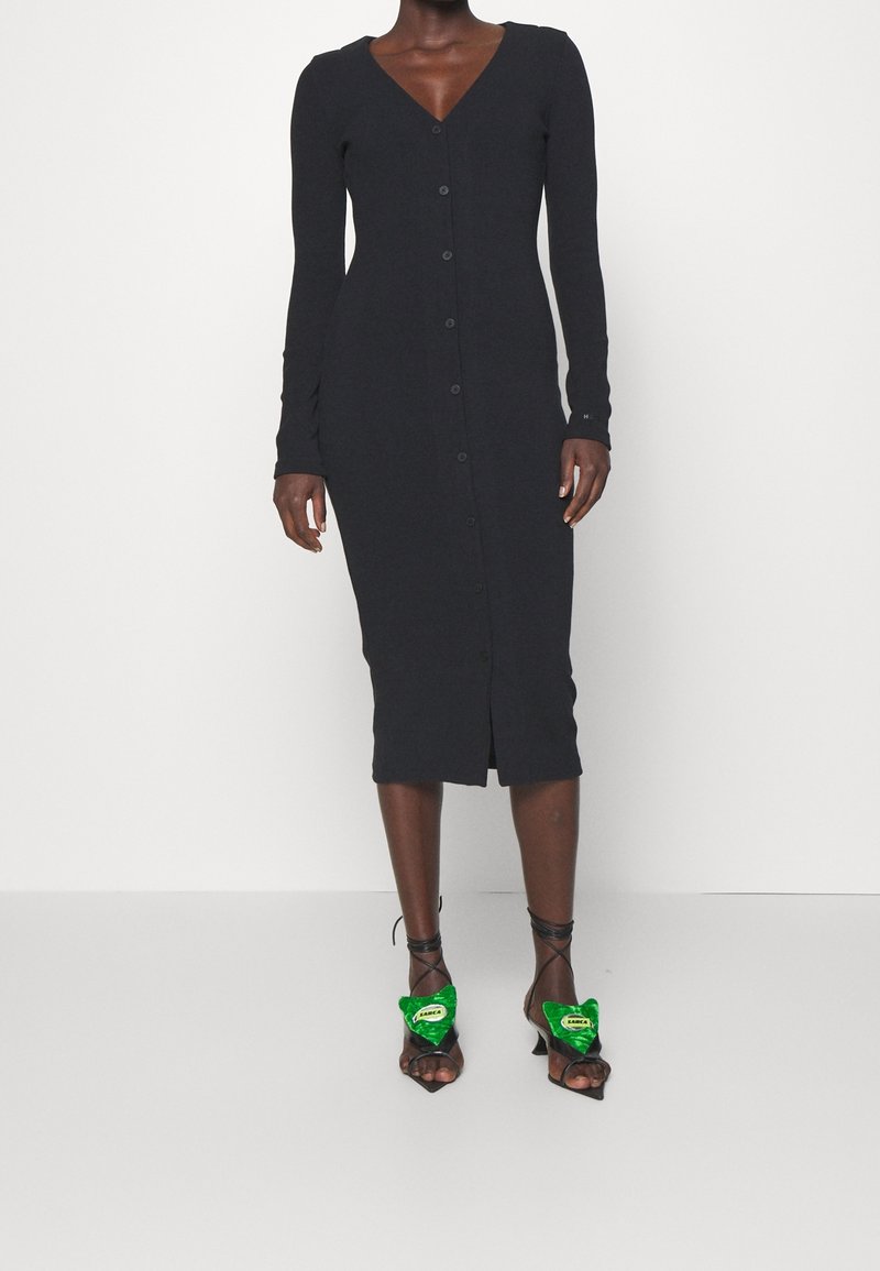 Femme portant une robe midi noire boutonnée et des chaussons verts en forme de cœur avec des brides noires à la cheville sur fond blanc.