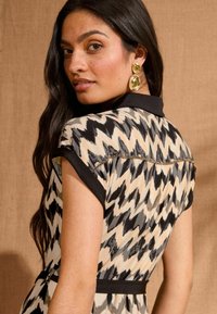 Rochie cu mânecă scurtă, cu un model în zigzag negru și bej, având o guler negru și un aplicație metalică pe cusătura umărului.